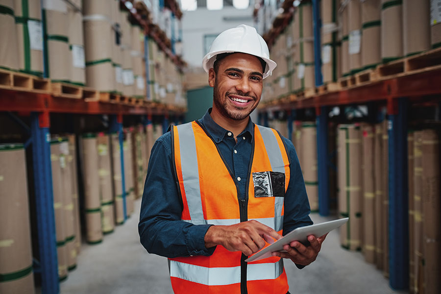Warehousing-and-Logistics-Insurance-Portrait-of-Smiling-Warehouse-Manager-Using-a-Digital-Tablet-in-a-Warehouse-While-Standing-between-Shelves-and-Looking-at-the-Camera